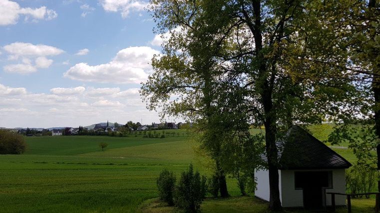 Eine ruhige Landschaft mit grünen Wiesen und einem kleinen Häuschen. Der Himmel ist teilweise bewölkt und es gibt einige Bäume in der Nähe.