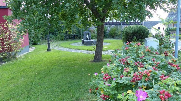 Ein gepflegter Garten mit einem großen Baum und einer Steinbank. Bunte Blumen blühen entlang des Weges.