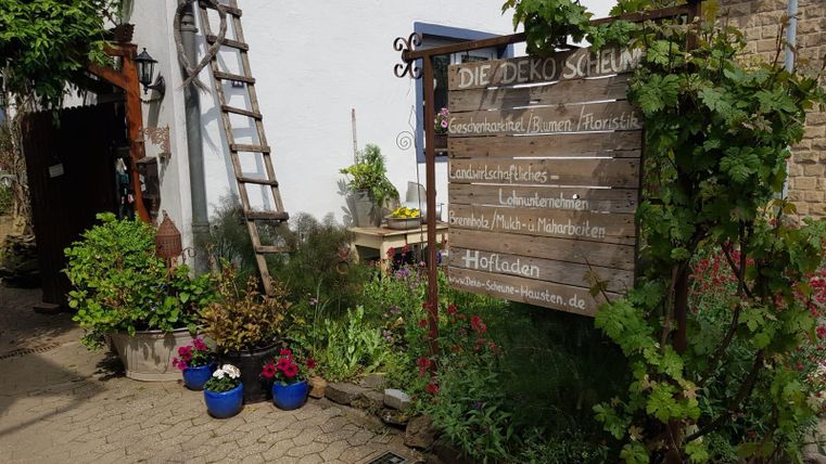 An inviting courtyard with a wooden sign that reads "THE DECOR BARN." Lush plants and colorful flower pots adorn the entrance area.