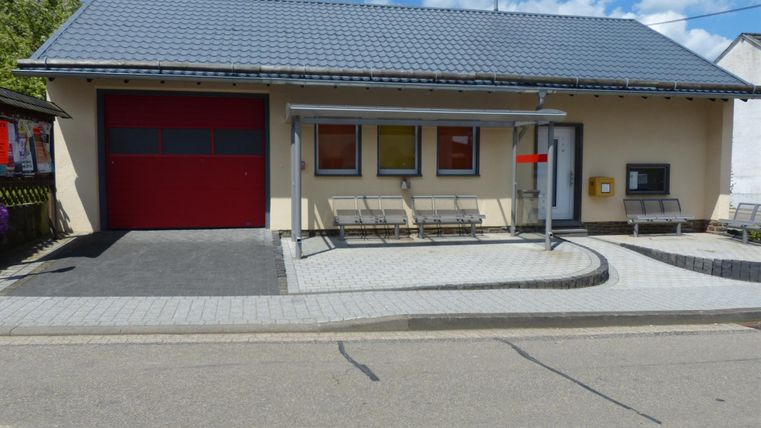 Ein einfaches Gebäude mit einem roten Garagentor und mehreren Fenstern. Vor dem Gebäude gibt es Sitzbänke und einen Haltestellenbereich.