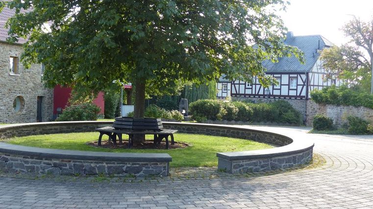 Ein ruhiger Platz mit einem großen Baum und einer Sitzgruppe aus Holz. Im Hintergrund sind traditionelle Häuser zu sehen.