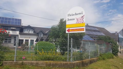 Ein Schild der Fleischerei Jonas steht vor einem Garten mit Blumen. Im Hintergrund sind Häuser und Solarpanels sichtbar.