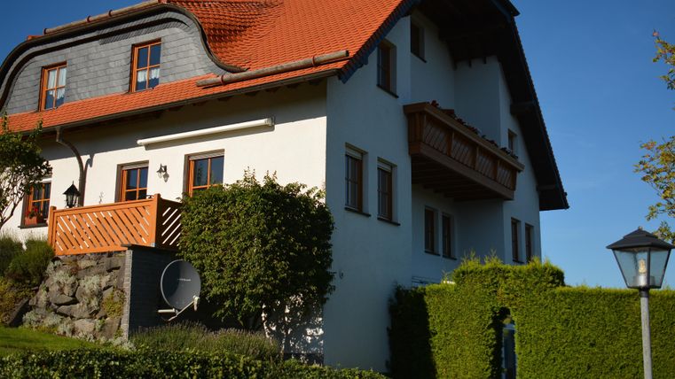 Ein modernes Haus mit rotem Dach und hellen Wänden. Es hat einen Balkon und einen gepflegten Garten.