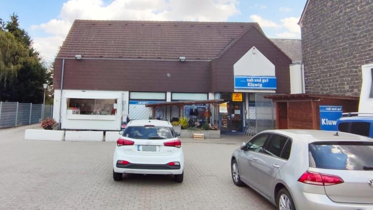 Ein Geschäft mit einem modernen Eingang und großen Fenstern. Vor dem Gebäude stehen mehrere Autos auf einem gepflasterten Parkplatz.