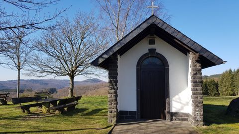 Eine kleine Kapelle mit einem dunklen Holzportal und einer Steinfassade. Im Hintergrund sind Bäume und sanfte Hügel zu sehen.