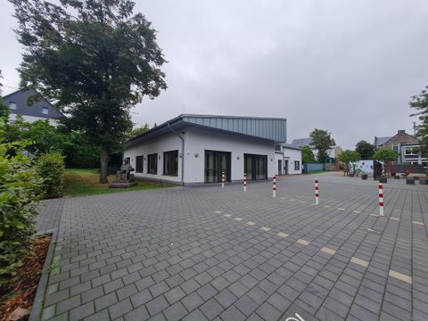 A modern building with large windows and a paved parking lot. Trees and other buildings are visible in the background.
