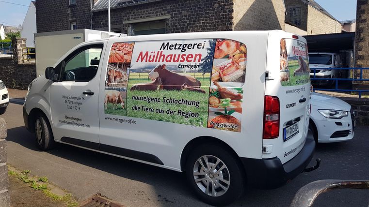 Ein Lieferwagen der Metzgerei Mühlhausen mit bunten Werbeaufdrucken. Er steht in einer engen Straße zwischen anderen Fahrzeugen.