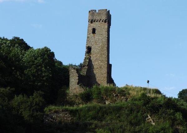 An old, stone tower stands on a hill, surrounded by trees and green vegetation. The sky is clear and blue.