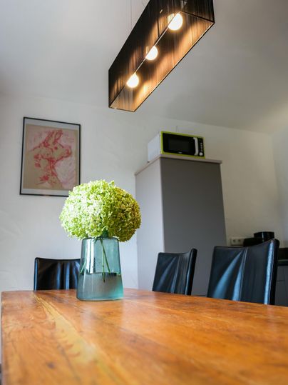 A cozy dining area with a wooden table and black chairs. In the center, there is a vase with a green flower and a modern pendant light above it.
