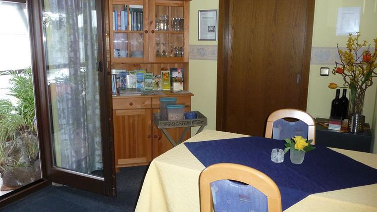 A cozy dining area with a table and wooden chairs. In the background, there is a bookshelf and a door to a balcony.