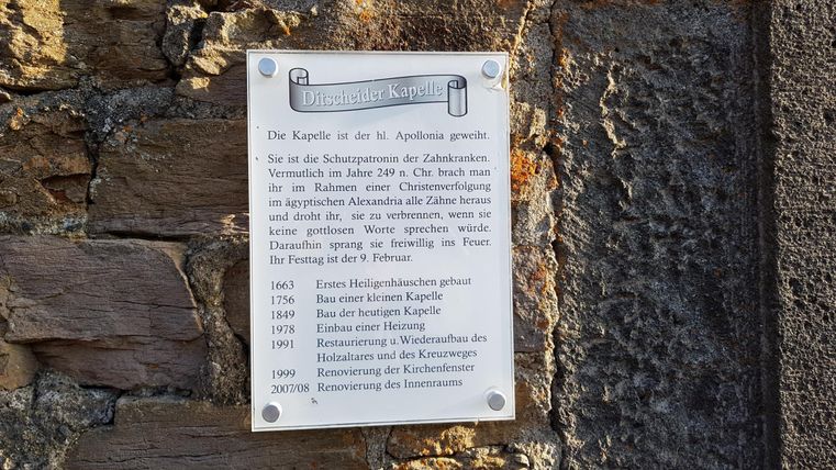 Eine Informationsplatte über die Kapelle zu St. Apollonia. Sie enthält historische Daten und Erklärungen zur Kapelle und ihrer Bedeutung.
