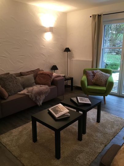 A cozy living room with a gray sofa and a green armchair. There are two small tables and large windows that let in light.