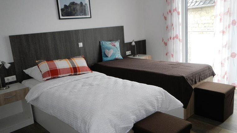 A bright bedroom with two single beds, a modern nightstand, and a beautiful view through large windows. The walls are adorned with light curtains and a decorative picture.