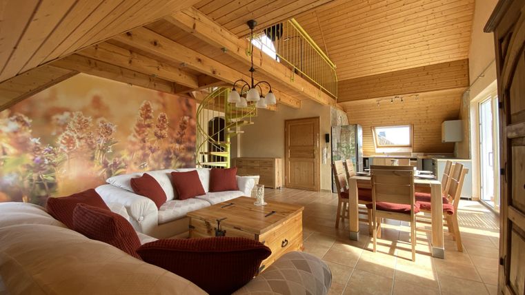 Ein gemütliches Wohnzimmer mit Holzdecke und großen Fenstern. Es gibt ein Sofa, einen Esstisch und eine warme Atmosphäre.