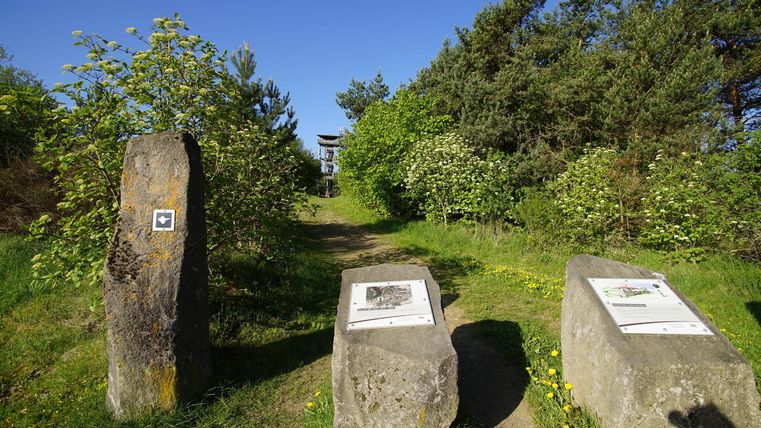 A path leads through a green environment with trees and bushes. Two large stones with information panels stand at the edge of the path.