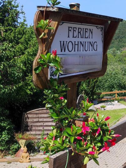 Ein Holzschild mit der Aufschrift "Ferienwohnung" steht umgeben von bunten Blumen. Im Hintergrund sind grüne Bäume und ein blauer Himmel zu sehen.