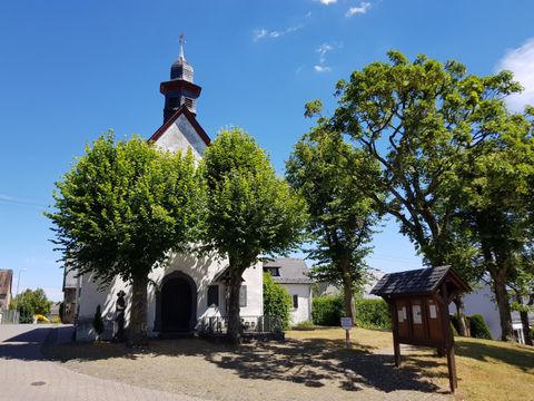 Eine kleine Kapelle mit einem Holzverkleidung und einem neugestalteten Dach. Umgeben von grünen Bäumen und blauem Himmel.