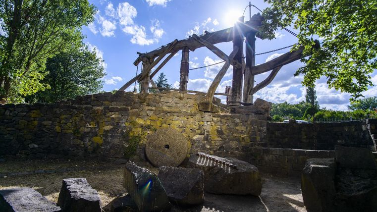 Alte Steinmühle mit Holzkonstruktion und Mühlsteinen im Freien, umgeben von Bäumen und blauem Himmel.