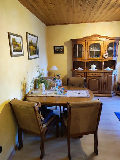 Ein gemütlicher Essbereich mit einem Holztisch und Stühlen. An der Wand hängen Bilder, und es gibt einen Schrank mit Geschirr.