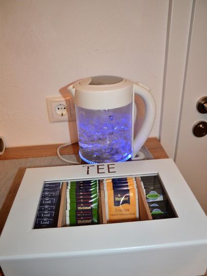 An electric kettle with blue light is standing on a table, while a tea box with various types of tea is lying next to it. The box is labeled "TEE."