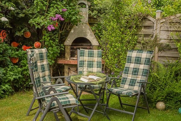 Ein gemütlicher Garten mit vier Stühlen und einem Tisch. Im Hintergrund steht ein Grill, umgeben von üppigem Grün und bunten Blumen.