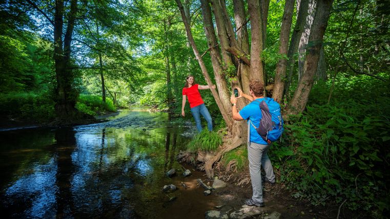Two people in a forest, one posing on a tree, the other taking a photo. A stream flows in the background.