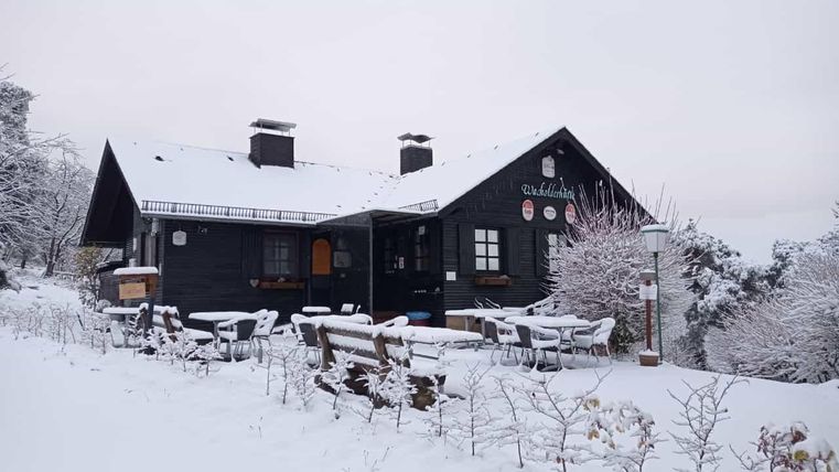 Ein gemütliches, schwarzes Haus im Schnee, umgeben von winterlichen Bäumen. Auf der Terrasse stehen Tische und Stühle, die mit Schnee bedeckt sind.