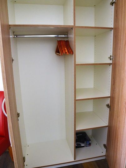 A empty, modern wardrobe with multiple compartments and a hanging rod area. Some wooden hangers are visible, and at the bottom there is a small safe.