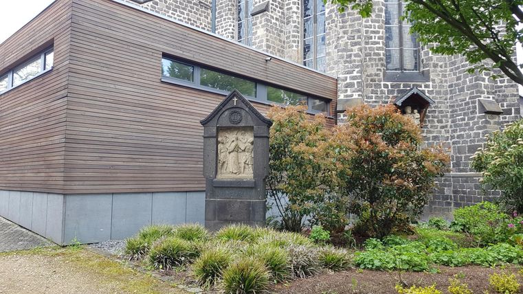 Ein modernes Gebäude aus Holz und Stein neben einer alten Kirche. Der Garten ist grün und gut gepflegt.