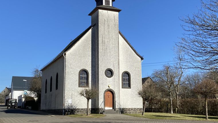 Eine kleine Kirche mit einem Glockenturm und einer schlichten Fassade. Der Himmel ist klar und blau, umgeben von ruhiger Landschaft.