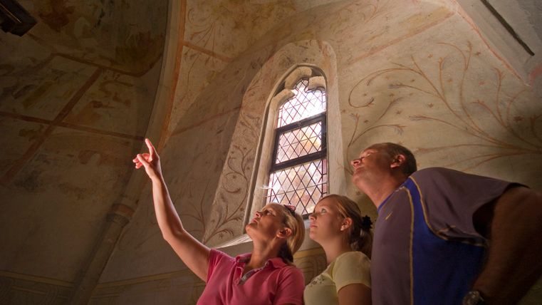 Three people look at frescoes in a church with a Gothic window.