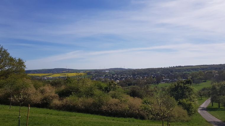 A picturesque landscape with gentle hills, trees, and a clear sky. In the background, small houses and fields are visible.