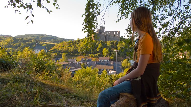 Person sitzt auf einem Baumstamm und blickt auf eine Burgruine in der Ferne.