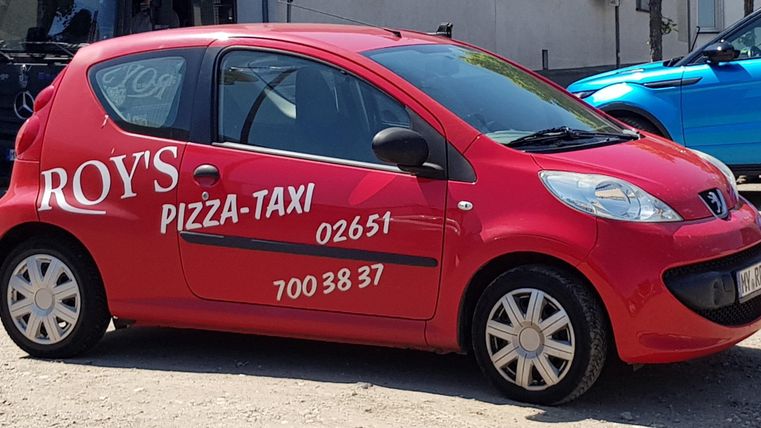 Ein rotes Taxi mit der Aufschrift "Roy's Pizza-Taxi". Das Fahrzeug steht auf einem Parkplatz und bietet einen klaren Blick auf die Türen.