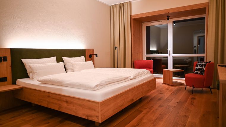 Ein modernes Hotelzimmer mit einem Doppelbett und Holzfußboden. Im Hintergrund gibt es große Fenster mit einem Ausblick und gemütliche Sitzmöbel.