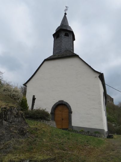 Eine kleine, weiße Kirche steht auf einem Hügel. Der Himmel ist bewölkt und die Umgebung ist grün.