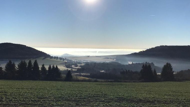 A vast landscape under a clear sky with the sun in the background. Fog lies over the hills and forests.