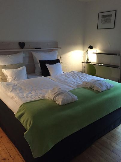 A cozy bedroom with a large bed and white bedding. A modern picture hangs on the wall, and there is a reading lamp as well as a shelf.
