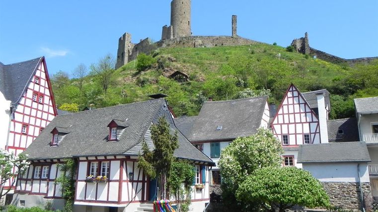 Eine malerische Landschaft mit Fachwerkhäusern im Vordergrund und einer historischen Burgruine auf einem Hügel im Hintergrund. Der Himmel ist klar und blau.