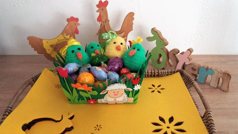 A colorful Easter nest with crocheted chicks and chocolate eggs. In the background, there is a wooden decoration with the inscription "Easter."
