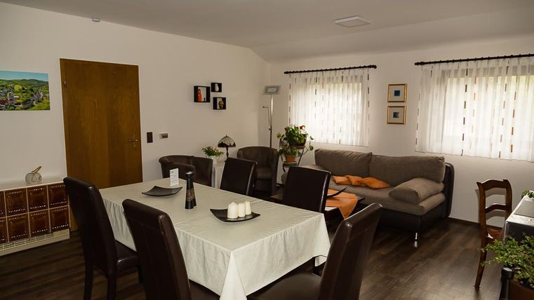 Ein gemütlicher Wohnbereich mit einem Esszimmertisch und Stühlen. Das Licht strömt durch die Fenster und es gibt eine Sofaecke mit Kissen.