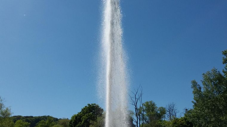 A water fountain gushes high into the blue sky. Surrounded by green trees and red stones, the scene is calming.