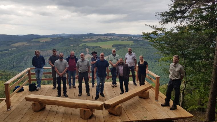 Eine Gruppe von Menschen steht auf einer Aussichtsplattform in der Natur. Im Hintergrund sind grüne Hügel und bewölkter Himmel zu sehen.