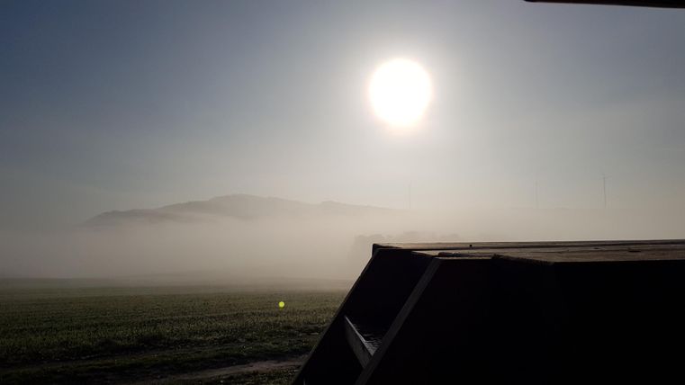 Eine neblige Landschaft mit der aufgehenden Sonne im Hintergrund. Im Vordergrund ist ein halbsichtbarer Gegenstand zu sehen.
