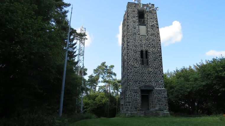 Ein alter Steinturm steht inmitten von Bäumen. Der Himmel ist blau mit einigen Wolken.