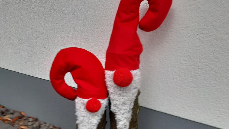 Two decorated tree trunks with red Christmas hats and a white beard. They are adorned with a red ribbon and stand in front of a white wall.