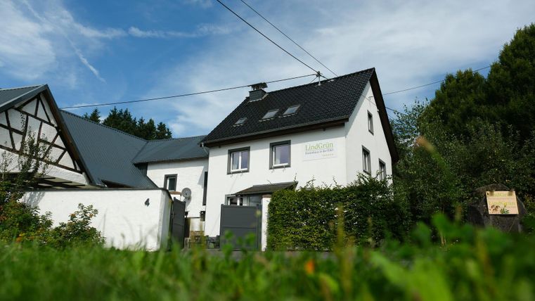 A modern house with a black roof and white walls. The garden is green and well-maintained, surrounded by trees and shrubs.