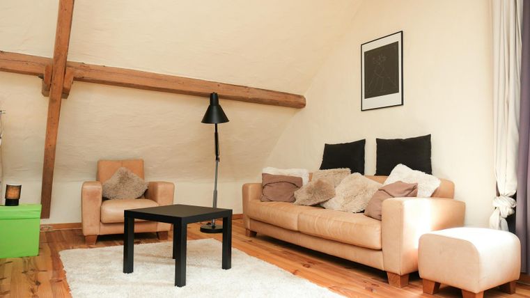 A cozy living room with a beige couch and an armchair. There is a carpet on the wooden floor, and a floor lamp stands next to the table.
