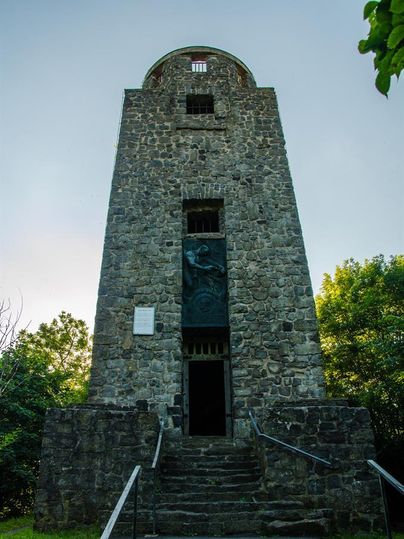 Ein steinerner Aussichtsturm mit einer Treppe, die zur Eingangstür führt. Umgeben von Bäumen und klarem Himmel.