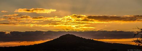 Ein malerischer Sonnenuntergang hinter einem Hügel. Die Wolken leuchten in warmen Gelb- und Orangetönen.